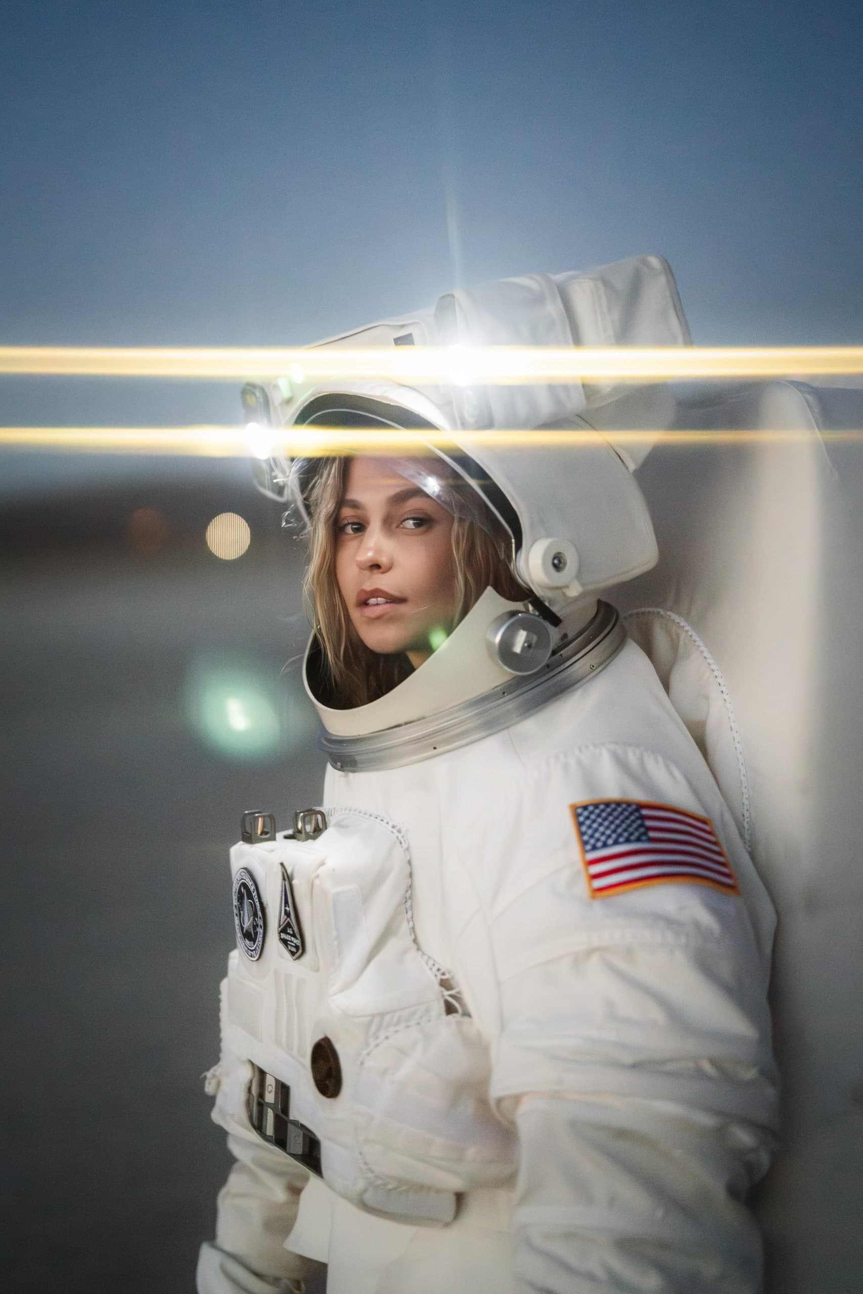 A woman in a full astronaut suit, dusk light, two bright horizontal light streaks crossing the frame.
