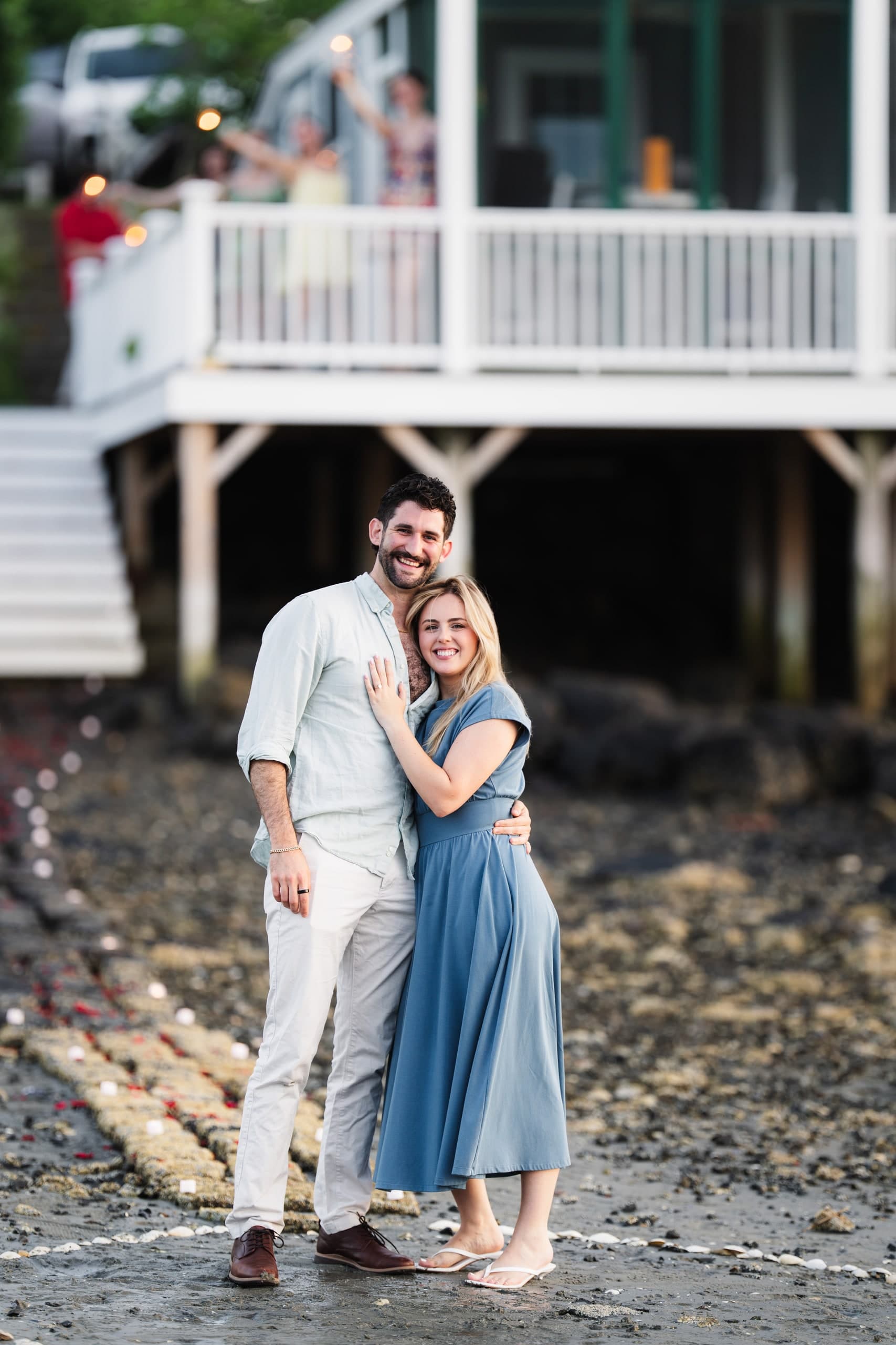 A couple standing on a rocky shoreline in front of a house with a porch, family visible above celebrating.