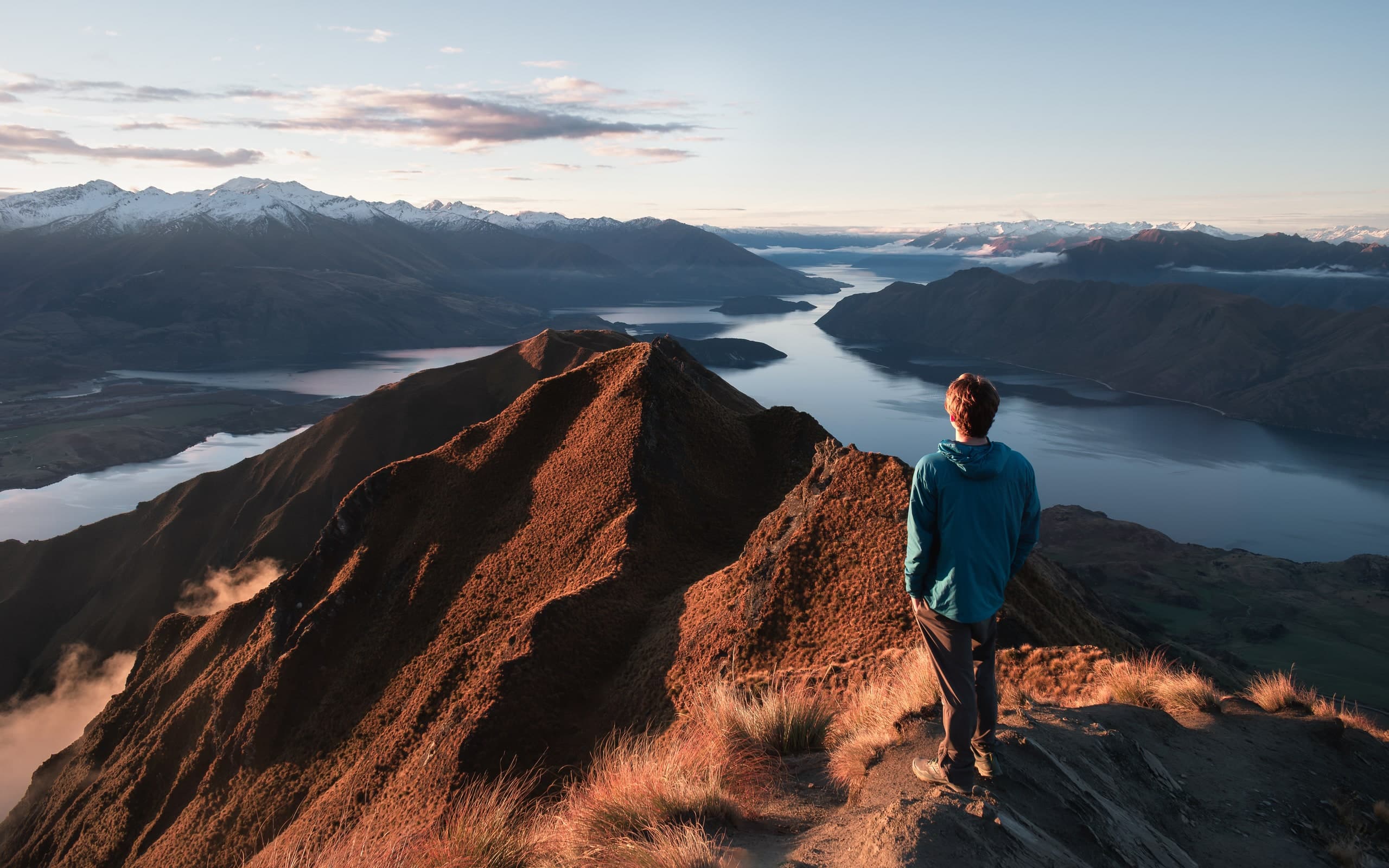 A hiker standing on the golden ridgeline of Roy's Peak above Lake Wanaka, New Zealand, blue mountains beyond.